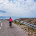 A girl in a sporty outfit, riding down a steep road on a bike. in the bottom Mediterranean Sea. Girl is enjoying the ride. Thick clouds above. Active holidays. Beautiful coastal line of Croatia